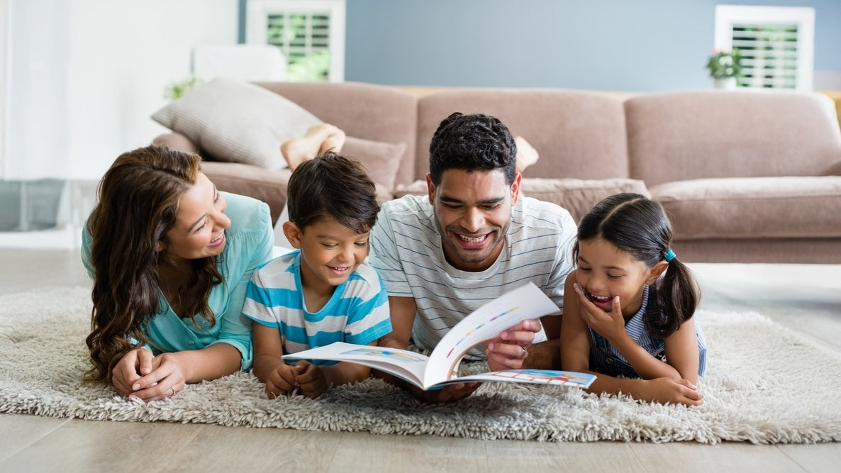 Family of four lying on the floor reading a book together in a living room.