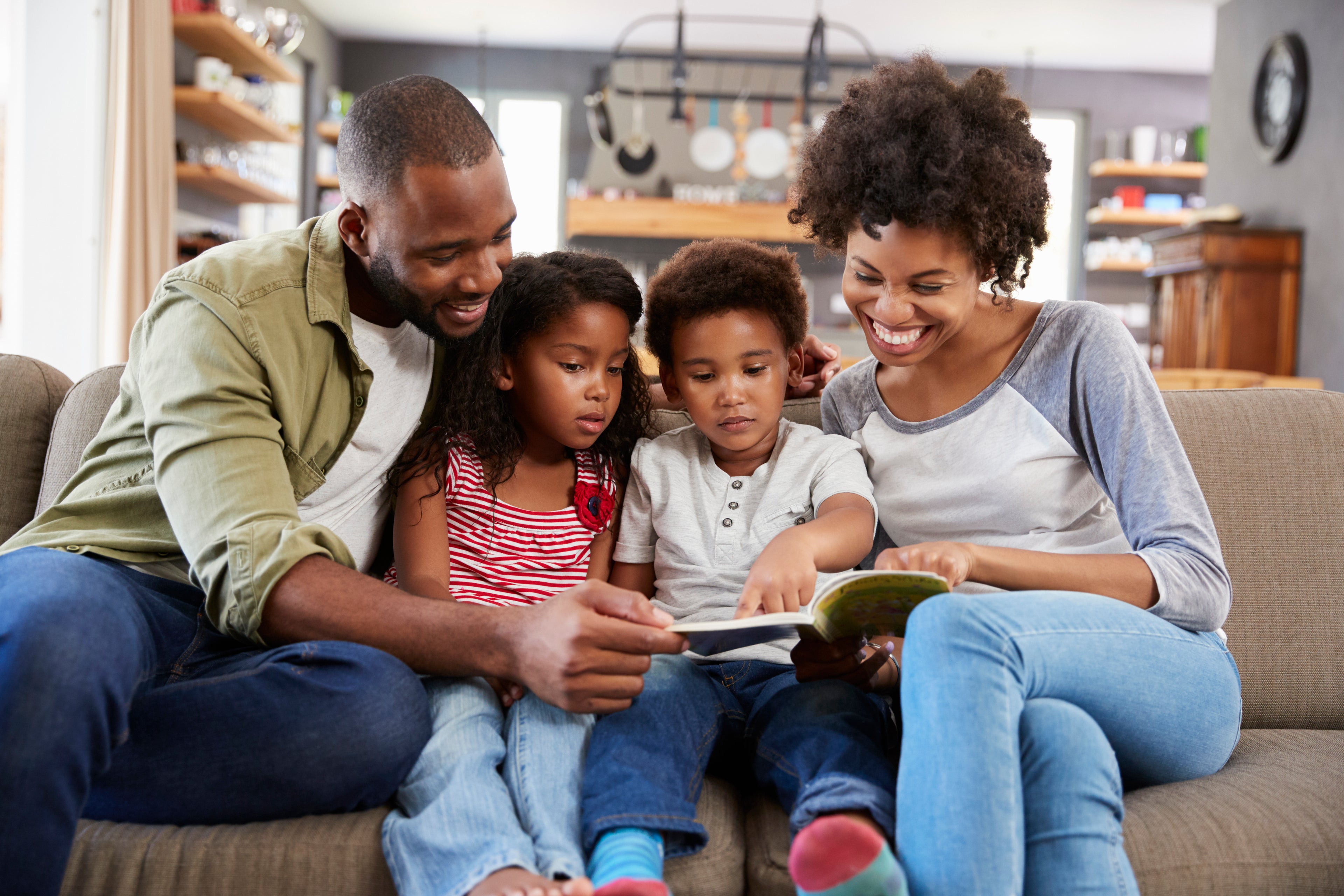 Family of four sitting on a couch reading a book together in a home setting.
